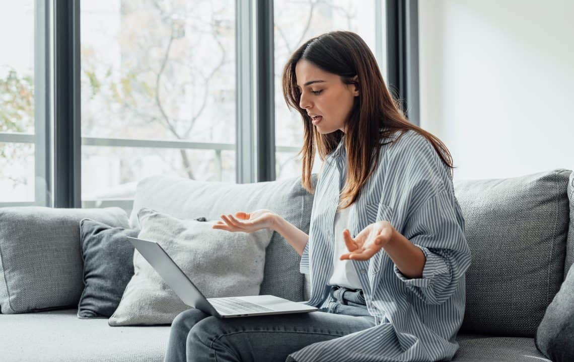 Angry young woman sit on sofa in living room frustrated having operational problems working on laptop, mad millennial girl feel stressed confused with slow internet connection or computer spam.